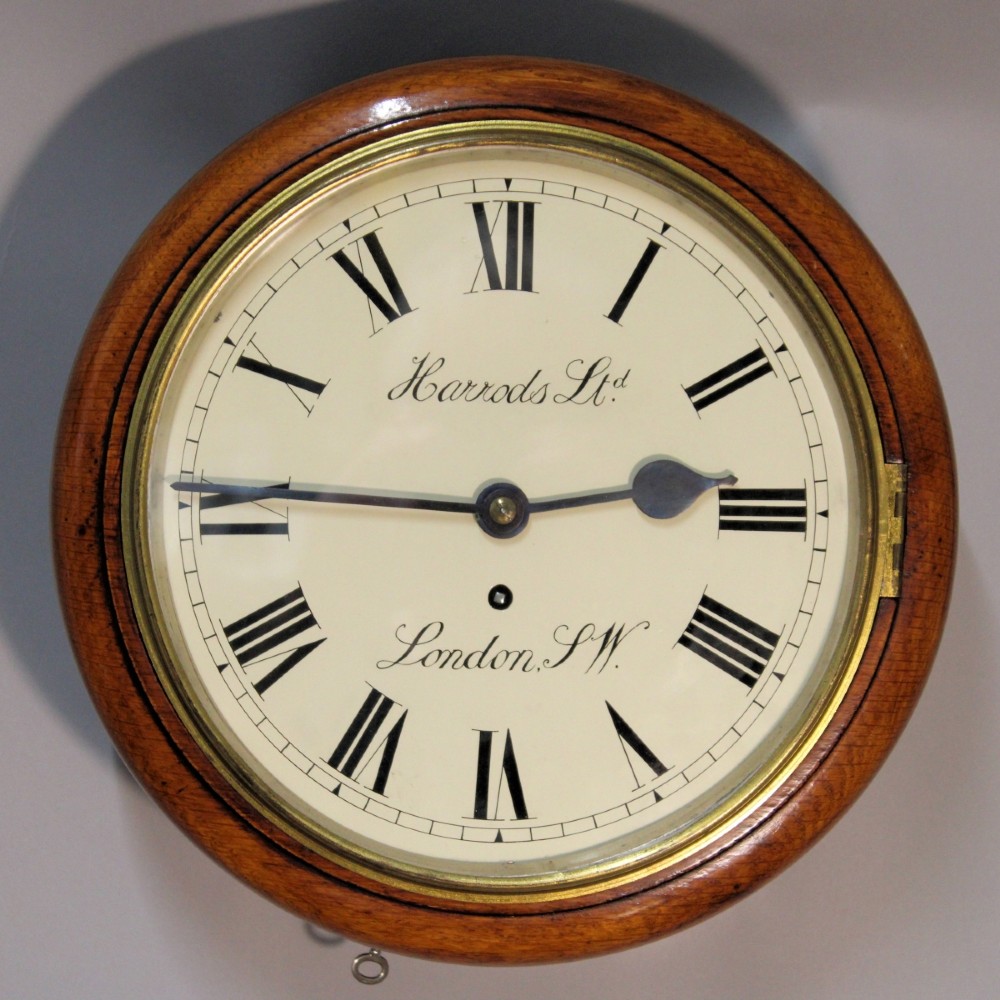 A circular Victorian oak fusee wall clock featuring a cream dial with black Roman numerals, brass bezel, and Harrods London retailer lettering.