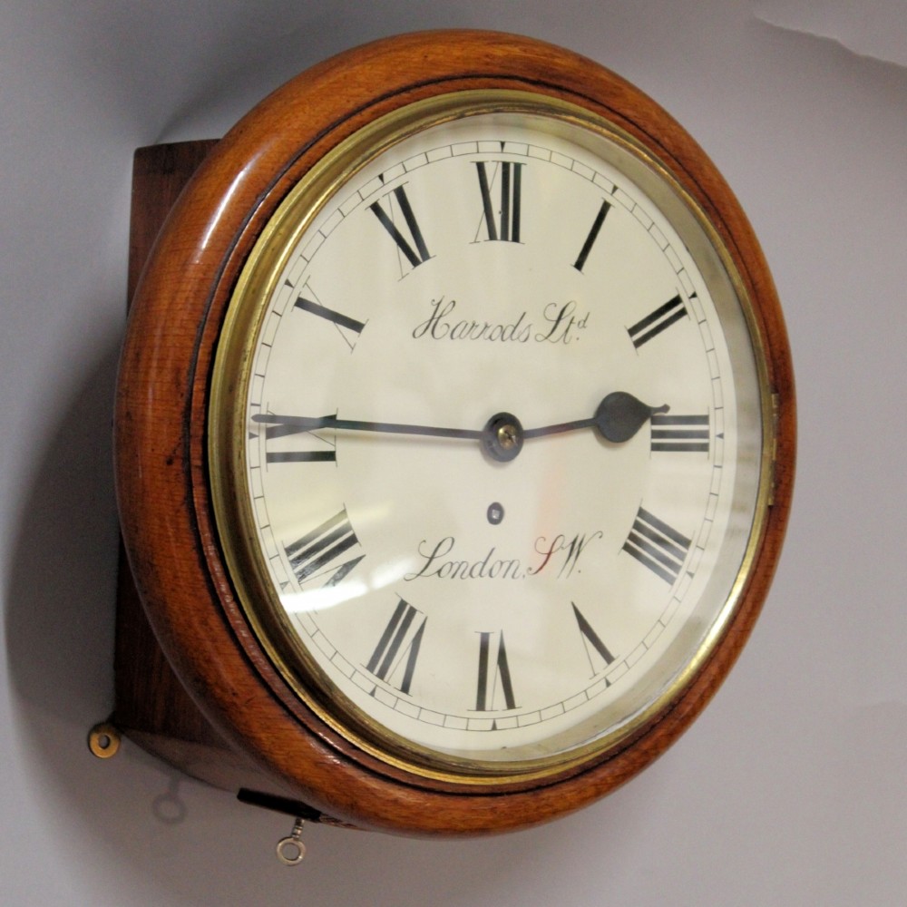 A circular Victorian oak fusee wall clock featuring a cream dial with black Roman numerals, brass bezel, and Harrods London retailer lettering. View II