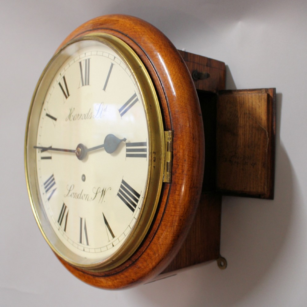 A circular Victorian oak fusee wall clock featuring a cream dial with black Roman numerals, brass bezel, and Harrods London retailer lettering. View III