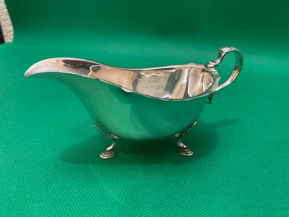 A polished silver sauce boat featuring a shaped rim, a scroll handle, and three pad feet.