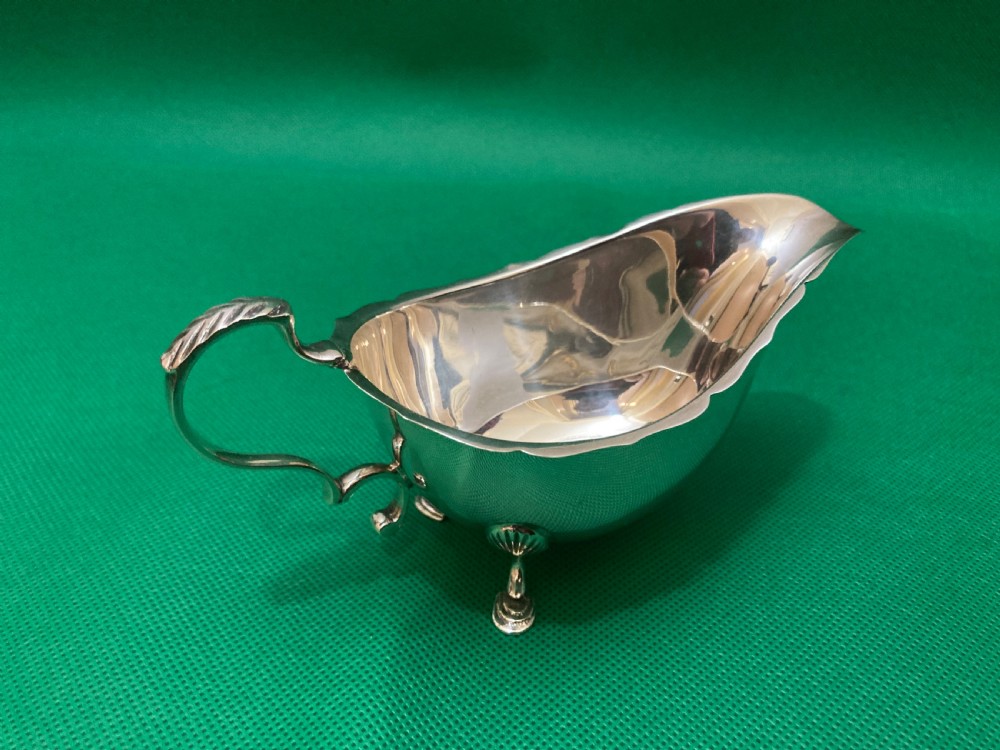 A polished silver sauce boat featuring a shaped rim, a scroll handle, and three pad feet. View II