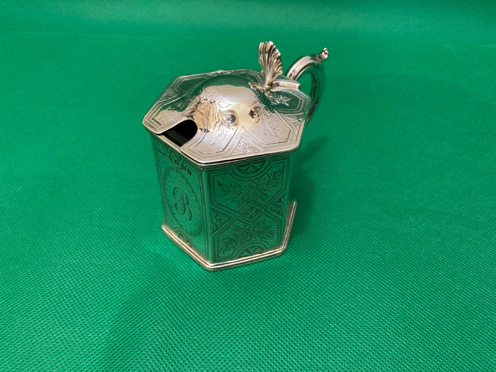 A Victorian-style hexagonal silver mustard pot with bright-cut foliate engraving, a shell thumbpiece, a curved handle, and a blue glass liner.