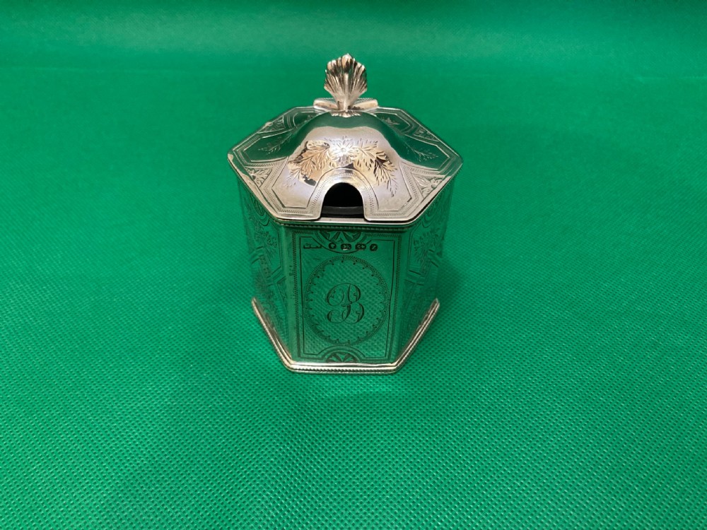 A Victorian-style hexagonal silver mustard pot with bright-cut foliate engraving, a shell thumbpiece, a curved handle, and a blue glass liner. View II
