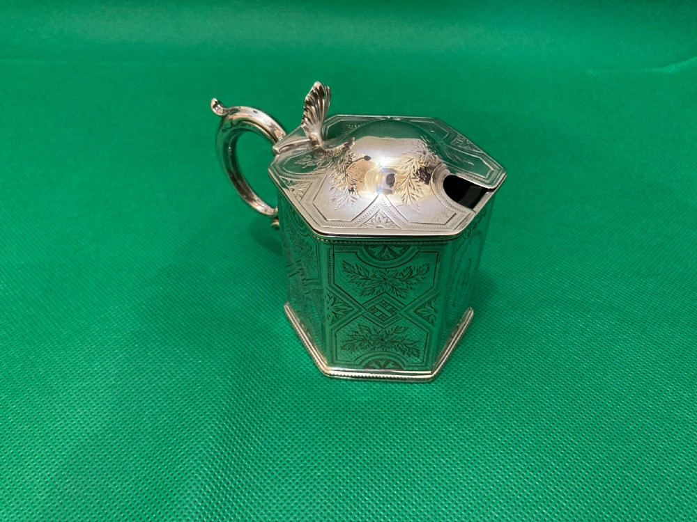 A Victorian-style hexagonal silver mustard pot with bright-cut foliate engraving, a shell thumbpiece, a curved handle, and a blue glass liner. View III