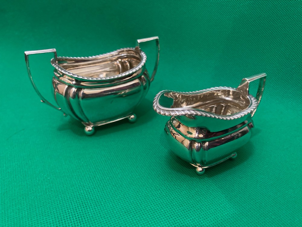 A matching silver cream jug and two-handled sugar bowl featuring gadrooned rims, angular handles, and small ball feet.