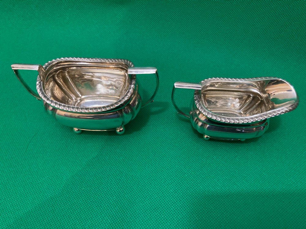 A matching silver cream jug and two-handled sugar bowl featuring gadrooned rims, angular handles, and small ball feet. View II