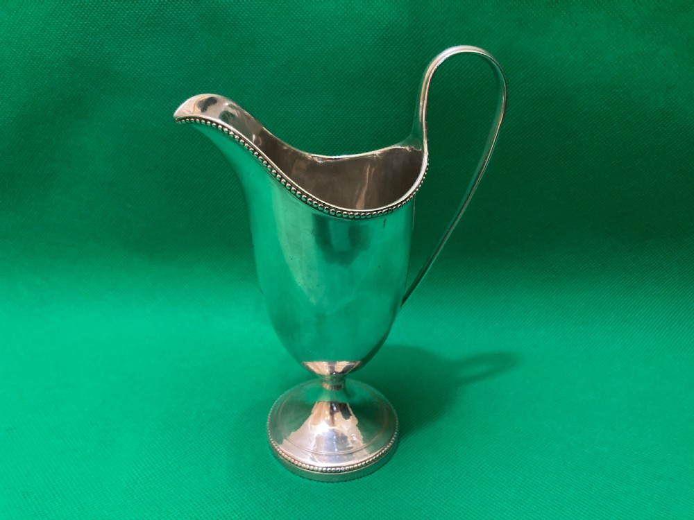 A polished silver helmet-shaped cream jug featuring a beaded rim, a high looped handle, and a round pedestal foot.