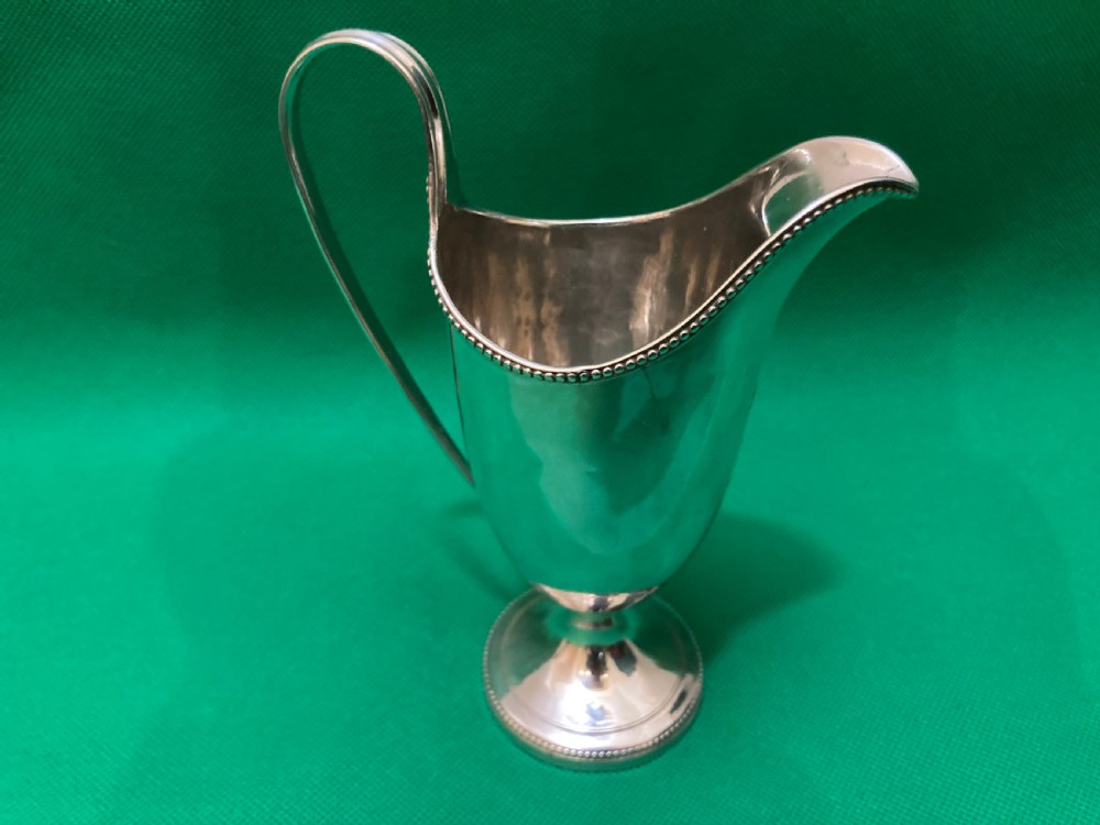 A polished silver helmet-shaped cream jug featuring a beaded rim, a high looped handle, and a round pedestal foot. View III