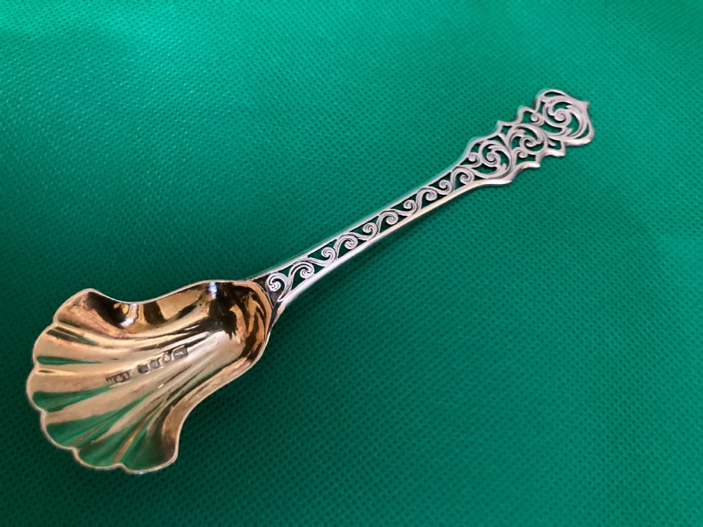 An antique silver sugar caddy spoon featuring a fluted shell-shaped bowl with British hallmarks and an openwork pierced scroll handle.