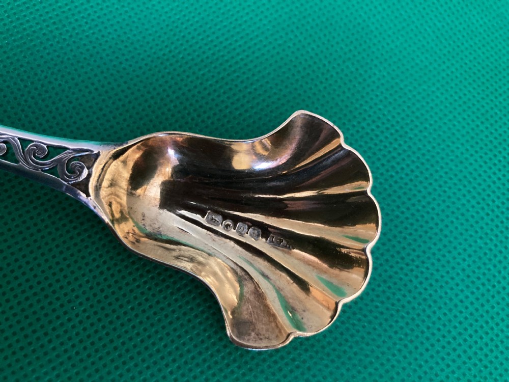 An antique silver sugar caddy spoon featuring a fluted shell-shaped bowl with British hallmarks and an openwork pierced scroll handle. View II
