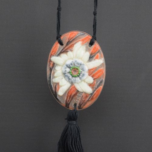 fig 1. An oval Art Deco pate-de-verre glass pendant featuring a white floral sea thistle motif surrounded by orange accents, suspended from a black cord with a lower thread tassel.