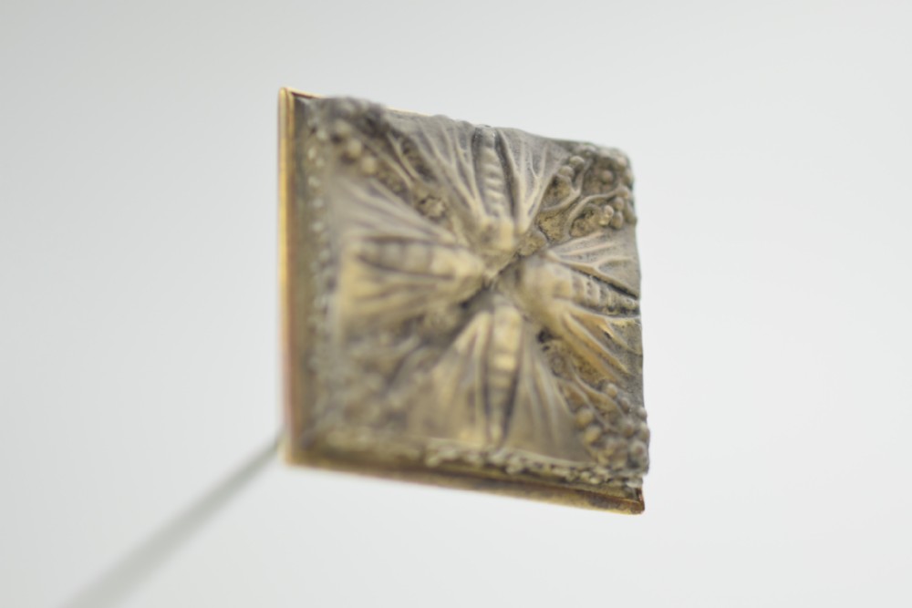 A square metal hatpin head featuring a relief motif of four butterflies meeting at the centre on a long pin shank.