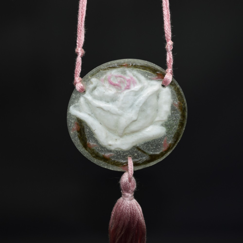 A circular translucent glass pate de verre pendant depicting a carved white rose with subtle pink highlights, suspended from a pink silk cord with a matching lower tassel.