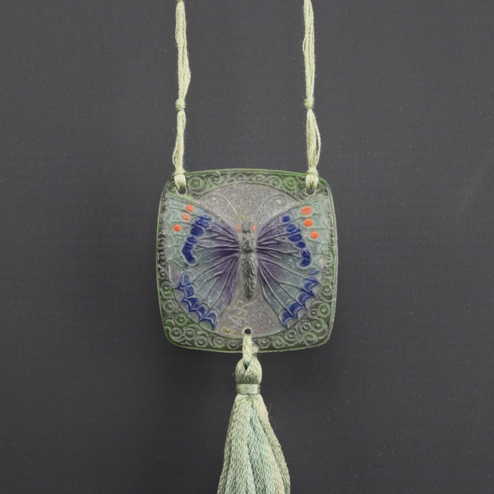 A square Art Deco pate de verre glass pendant featuring a relief butterfly motif in purple and blue against a translucent ground, suspended from a silk cord with a matching tassel.