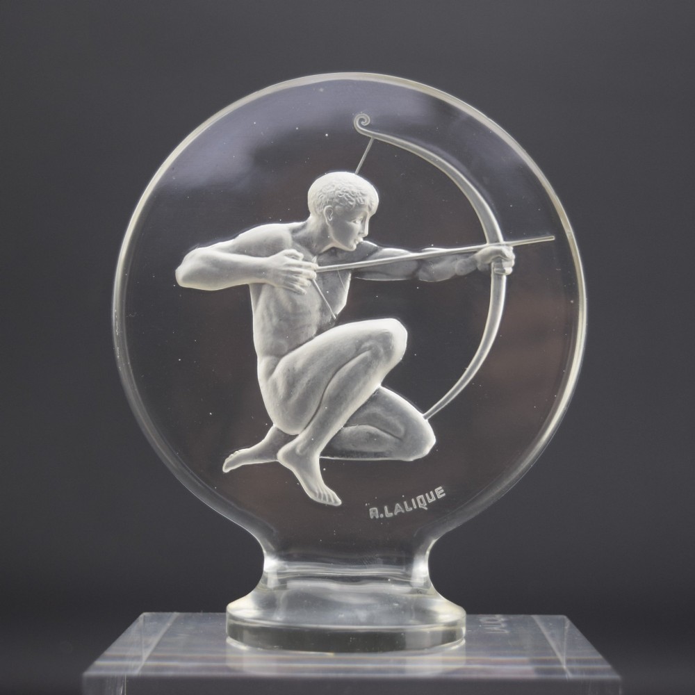 A circular clear and frosted Art Deco glass car mascot by Rene Lalique featuring a relief carving of a kneeling archer drawing a bow.
