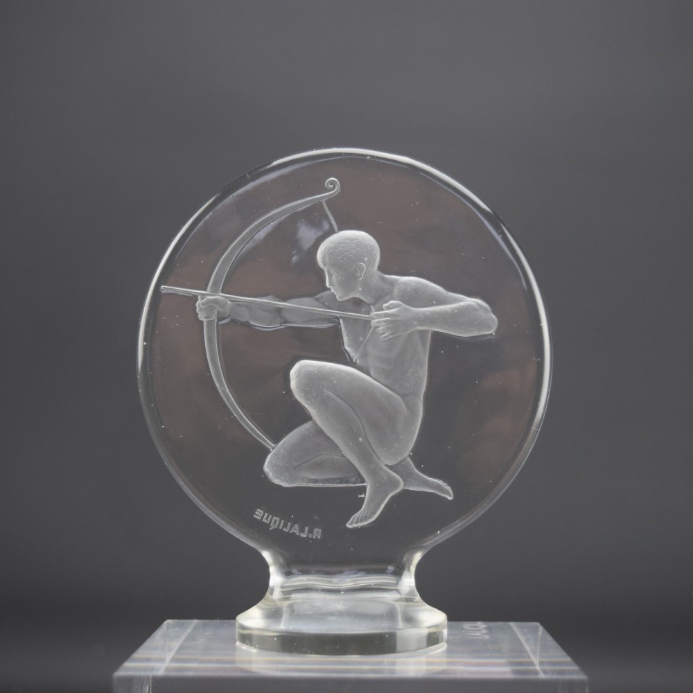A circular clear and frosted Art Deco glass car mascot by Rene Lalique featuring a relief carving of a kneeling archer drawing a bow. View IV