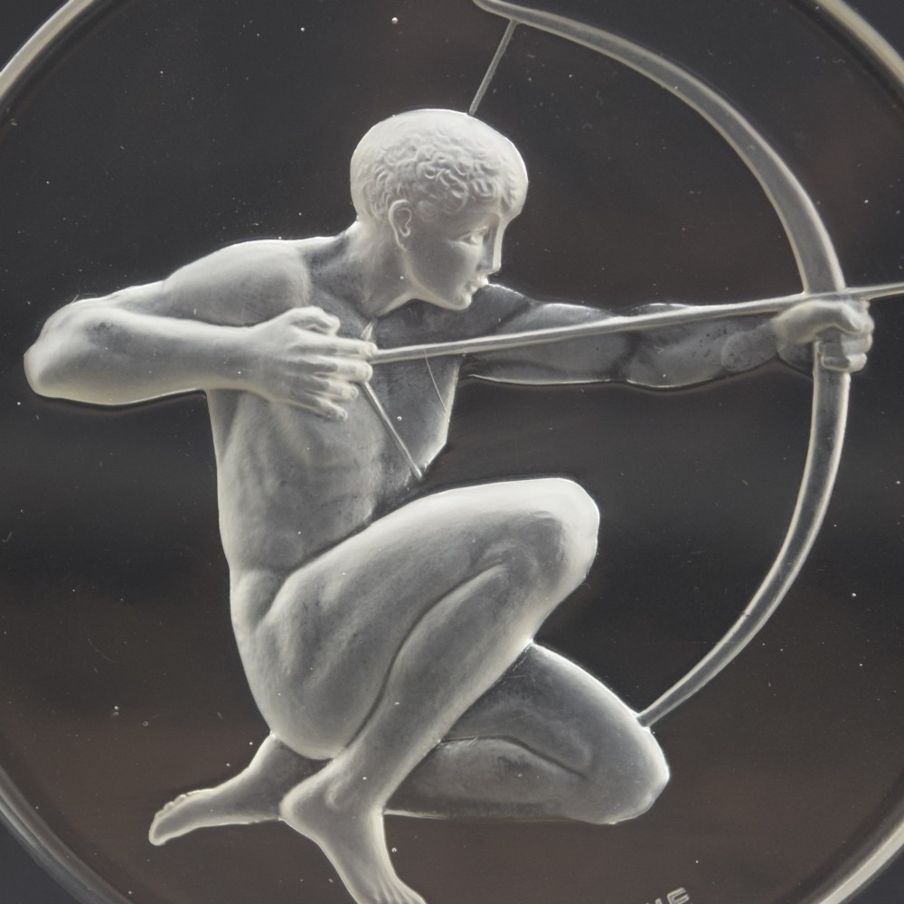 A circular clear and frosted Art Deco glass car mascot by Rene Lalique featuring a relief carving of a kneeling archer drawing a bow. View VII
