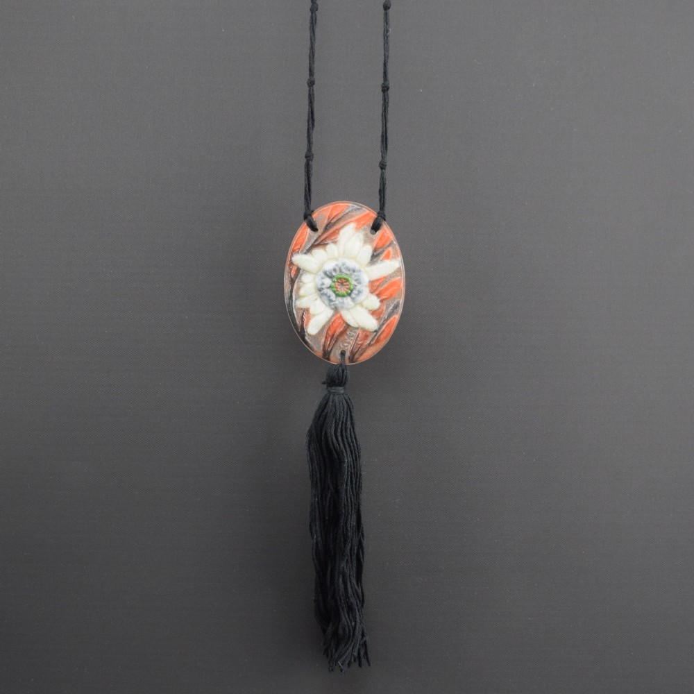 An oval Art Deco pate-de-verre glass pendant featuring a white floral sea thistle motif surrounded by orange accents, suspended from a black cord with a lower thread tassel.