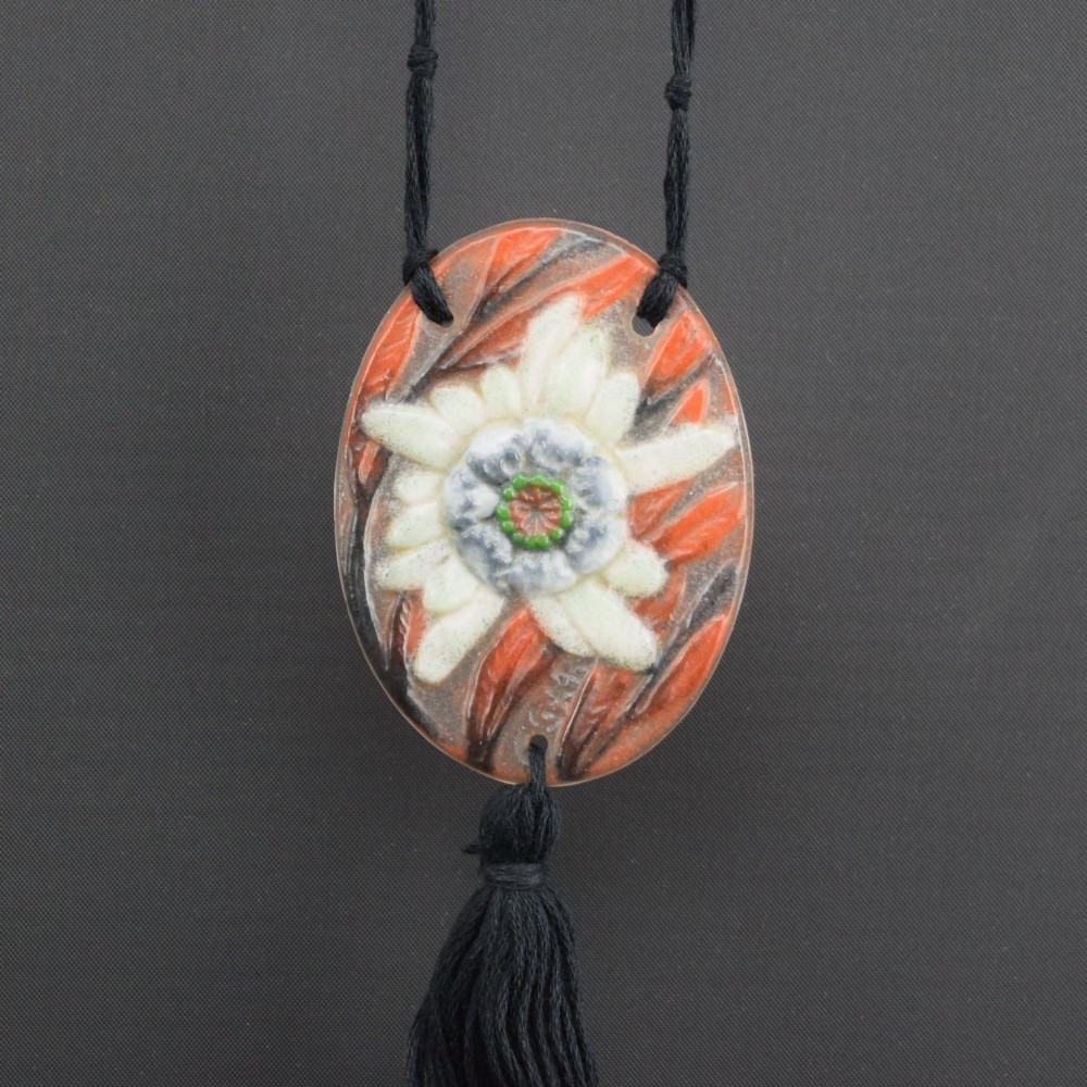 An oval Art Deco pate-de-verre glass pendant featuring a white floral sea thistle motif surrounded by orange accents, suspended from a black cord with a lower thread tassel. View II