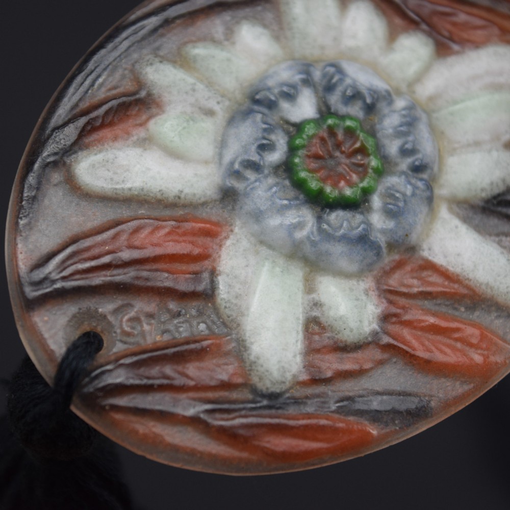 An oval Art Deco pate-de-verre glass pendant featuring a white floral sea thistle motif surrounded by orange accents, suspended from a black cord with a lower thread tassel. View IV