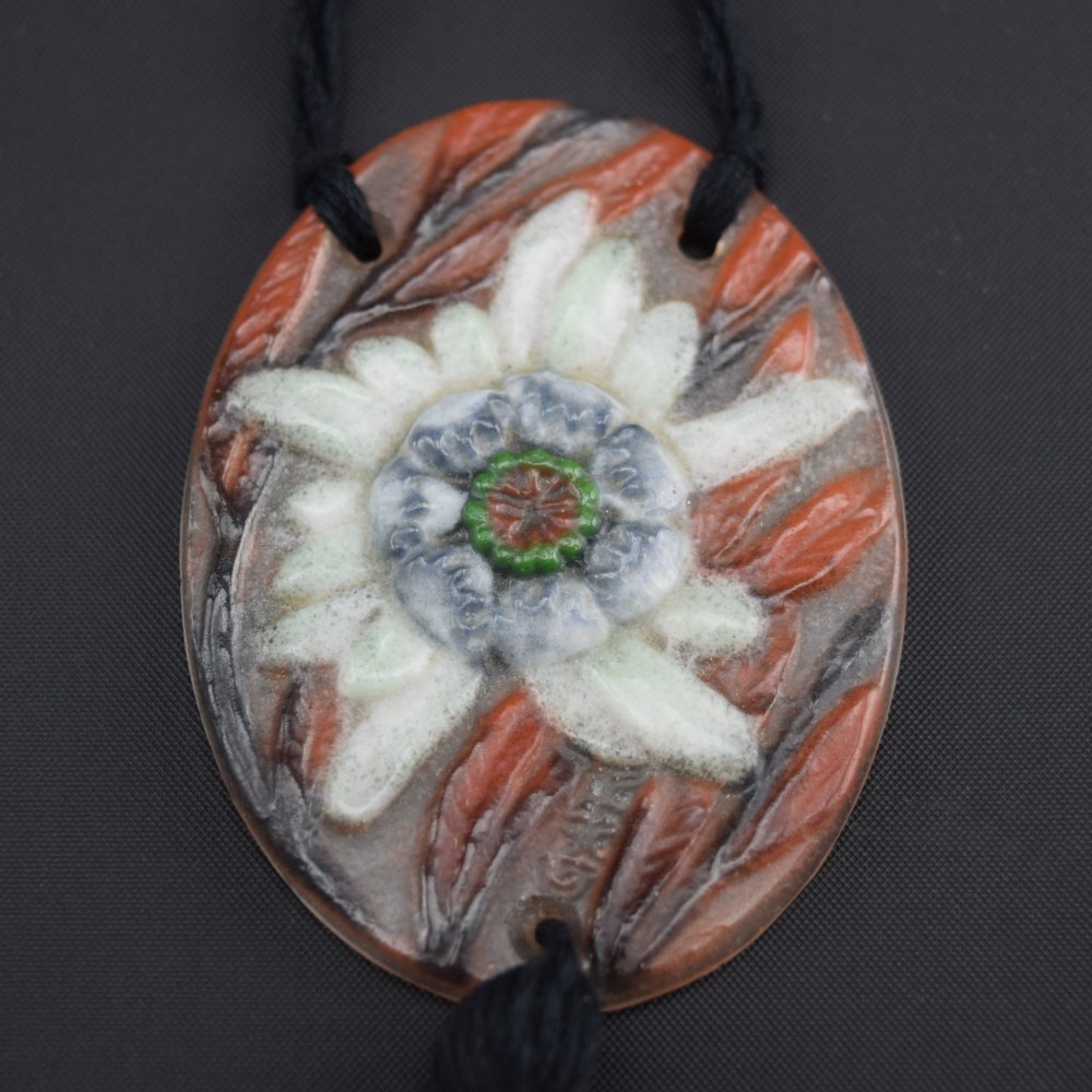 An oval Art Deco pate-de-verre glass pendant featuring a white floral sea thistle motif surrounded by orange accents, suspended from a black cord with a lower thread tassel. View V