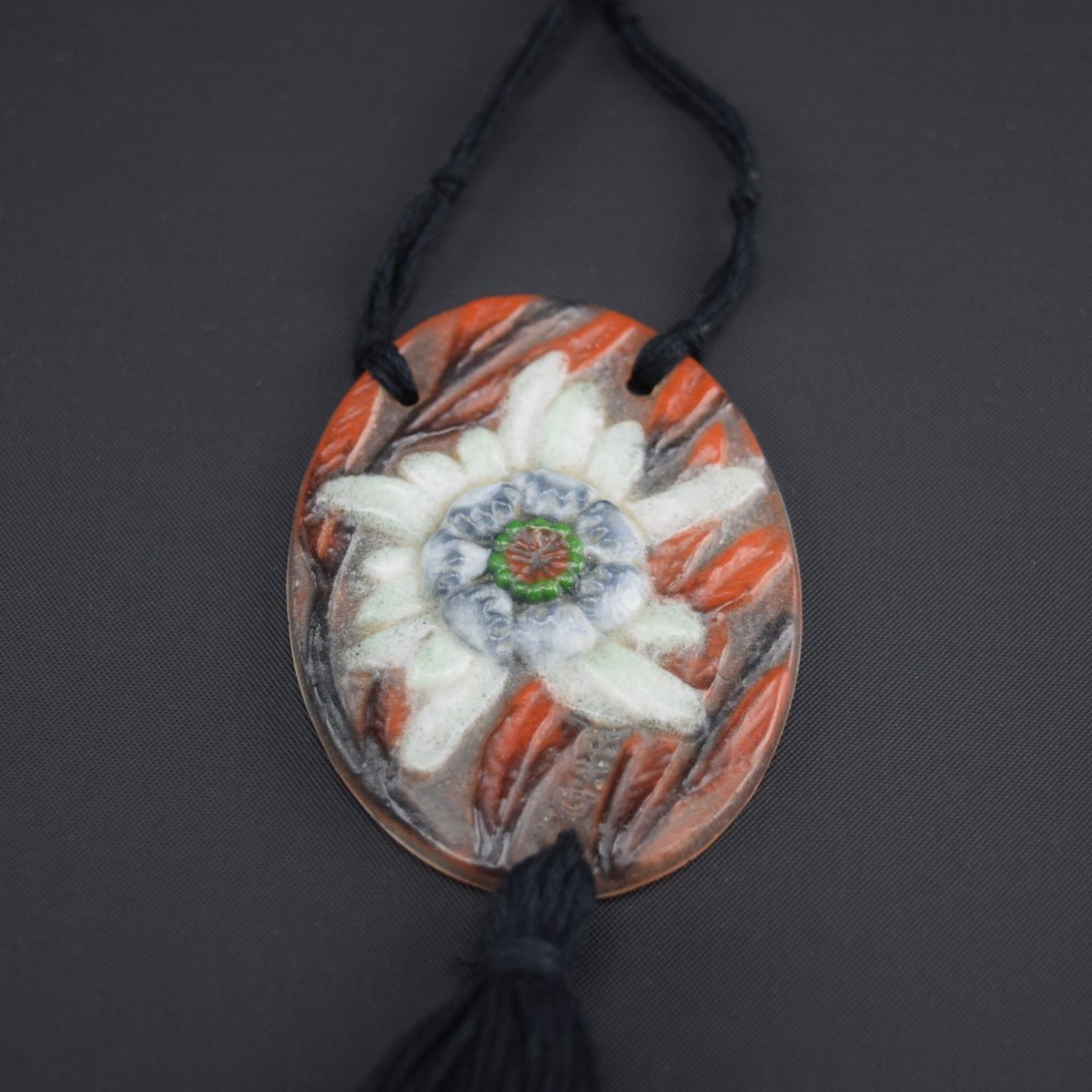 An oval Art Deco pate-de-verre glass pendant featuring a white floral sea thistle motif surrounded by orange accents, suspended from a black cord with a lower thread tassel. View VII