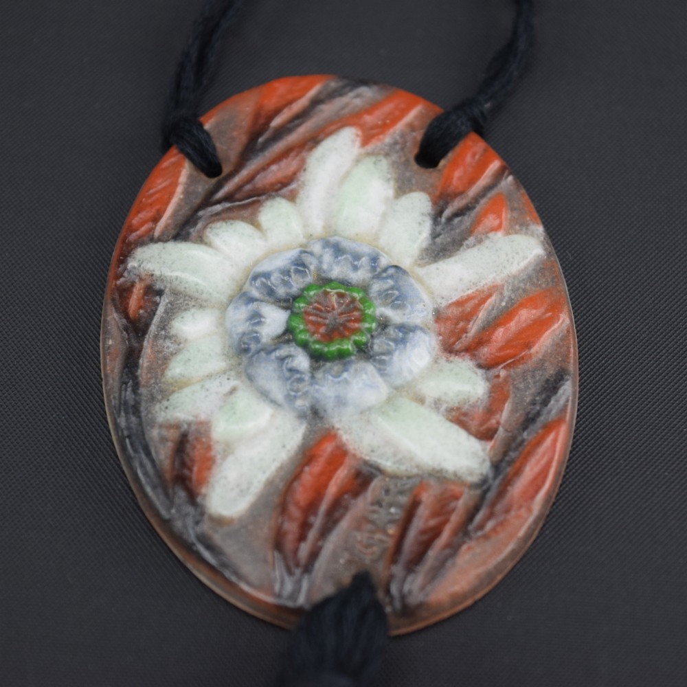 An oval Art Deco pate-de-verre glass pendant featuring a white floral sea thistle motif surrounded by orange accents, suspended from a black cord with a lower thread tassel. View VIII