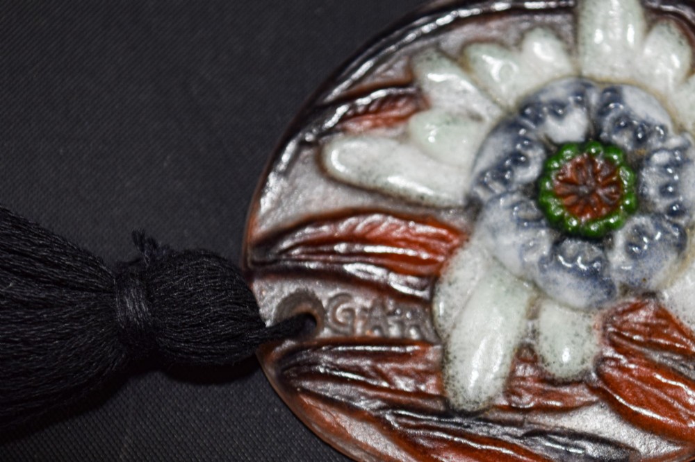 An oval Art Deco pate-de-verre glass pendant featuring a white floral sea thistle motif surrounded by orange accents, suspended from a black cord with a lower thread tassel. View IX