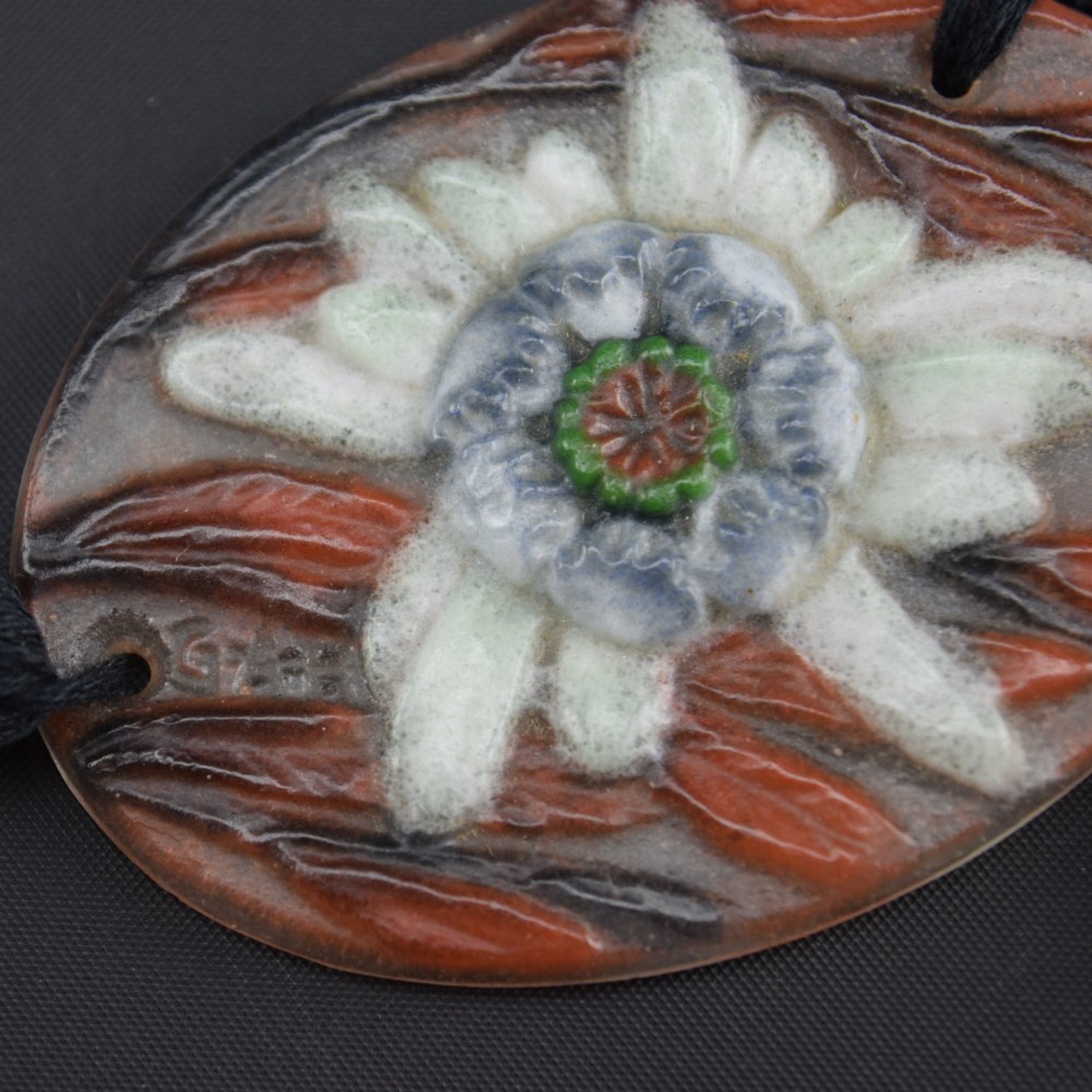 An oval Art Deco pate-de-verre glass pendant featuring a white floral sea thistle motif surrounded by orange accents, suspended from a black cord with a lower thread tassel. View X