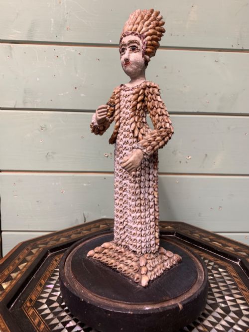 fig 1. A standing figure of a woman sculpted from tiny arranged seashells on a circular dark wood base.