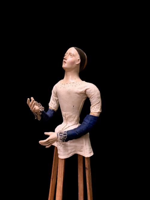 fig 1. A 19th-century processional mannequin with carved polychrome head and hands, dressed in cream fabric with dark blue sleeves, standing on a four-legged wooden cage base.