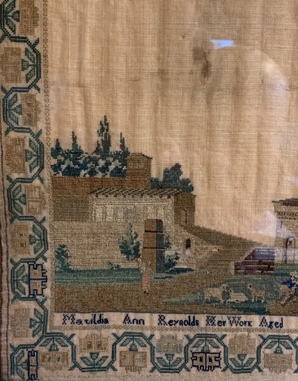A framed early nineteenth-century needlework sampler featuring an embroidered architectural scene and floral border on a natural linen ground. View V