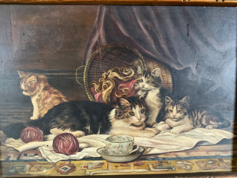 A 19th-century oil painting on board featuring a cat and three kittens beside a teacup, yarn balls, and a woven basket inside a wooden frame.