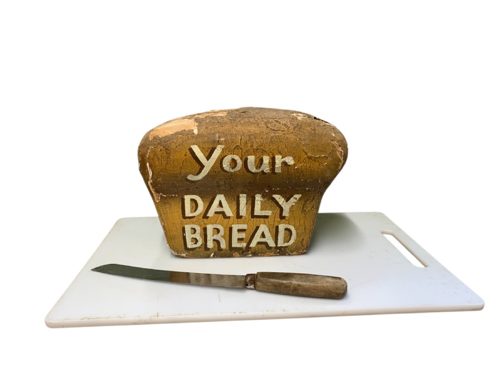 A vintage painted display model of a loaf of bread featuring hand-lettered text reading Your Daily Bread, presented alongside a wooden-handled knife. View III