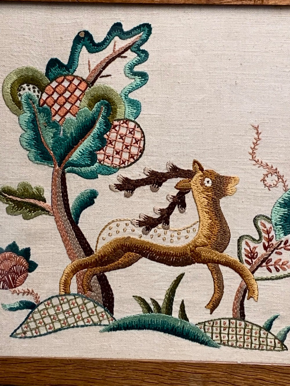 A square wooden frame enclosing a 1930s crewelwork embroidery depicting a leaping stag beside a stylised tree on a cream linen background. View IV