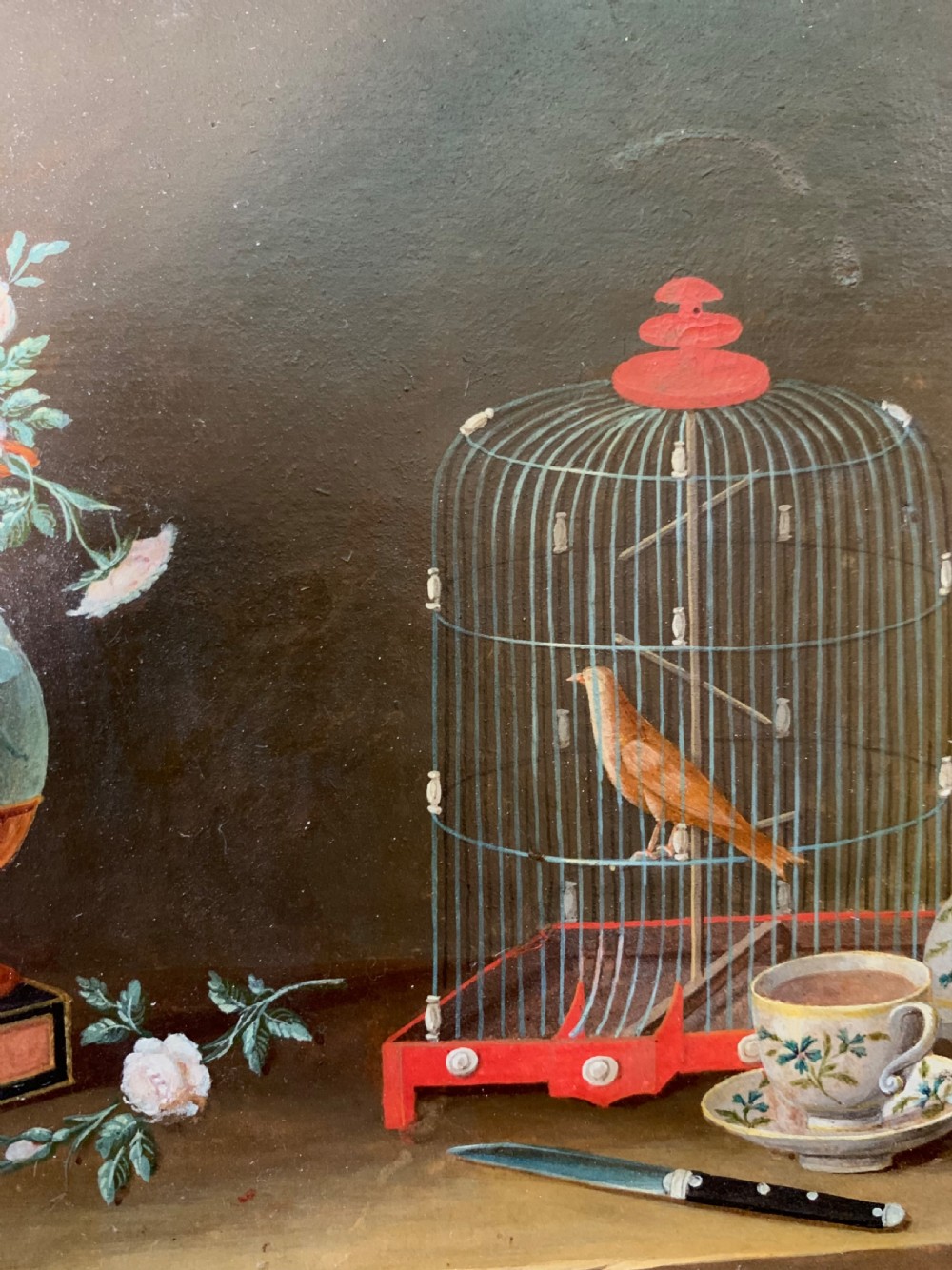 An early nineteenth-century framed French gouache still-life painting depicting a tabletop scene with a bird in a cage, a floral vase, porcelain teaware, and a book set within a carved giltwood frame. View VI