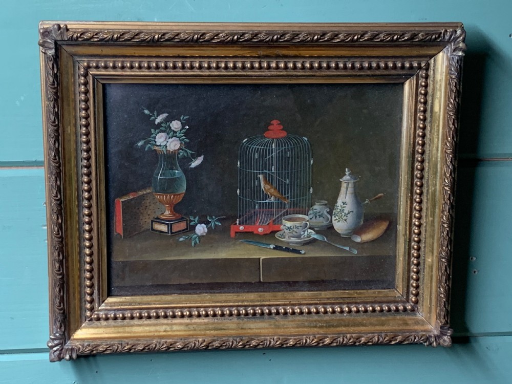 An early nineteenth-century framed French gouache still-life painting depicting a tabletop scene with a bird in a cage, a floral vase, porcelain teaware, and a book set within a carved giltwood frame. View VIII