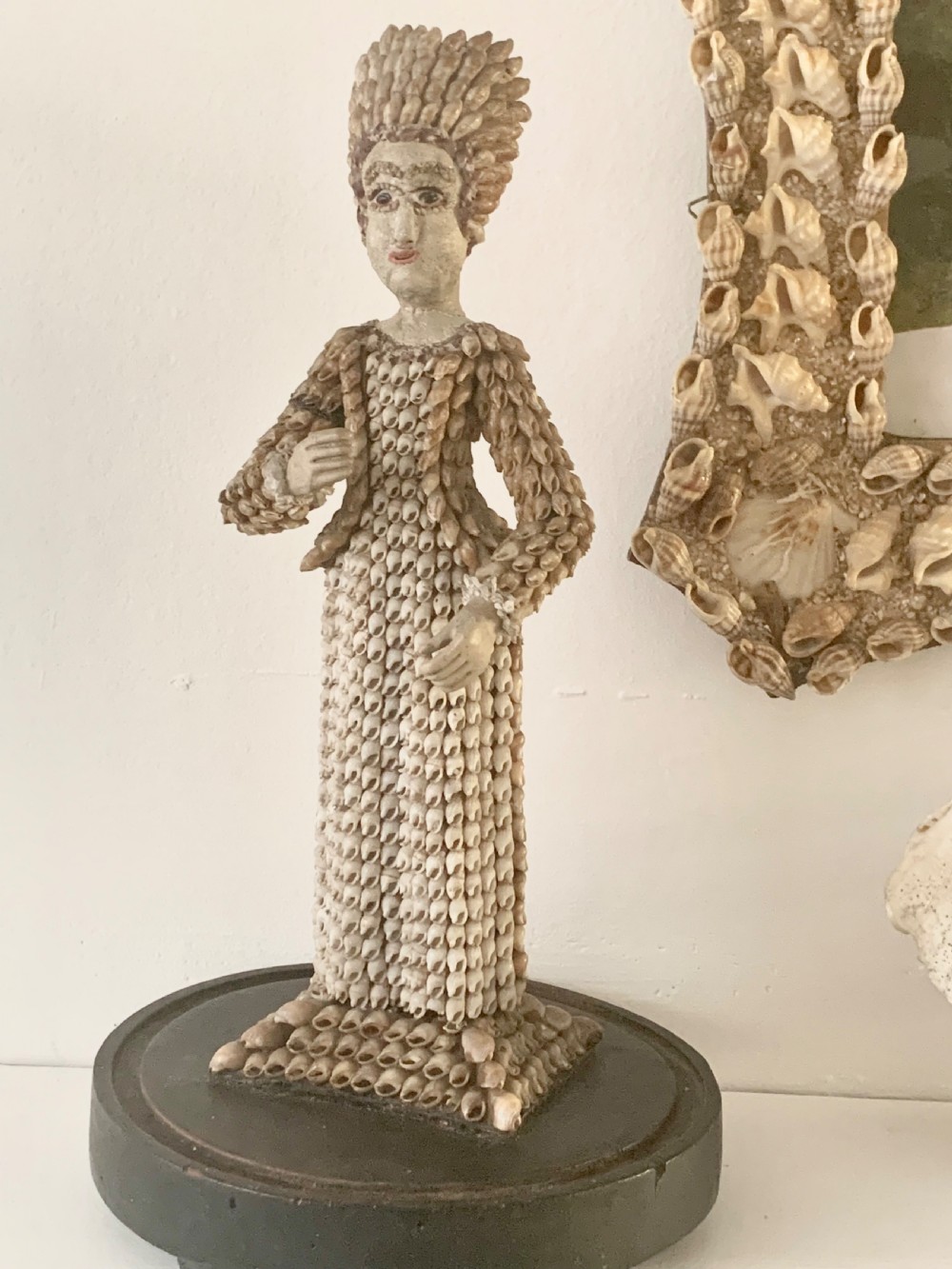 A standing figure of a woman sculpted from tiny arranged seashells on a circular dark wood base.