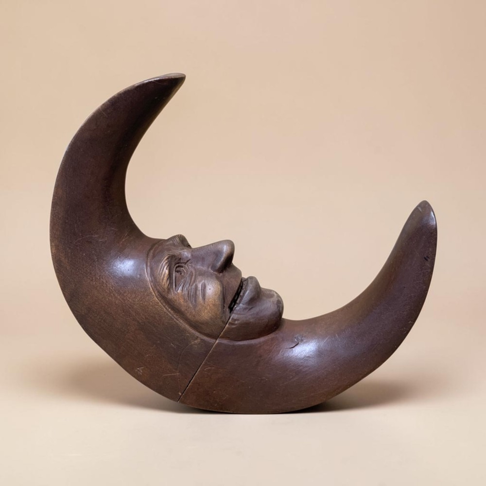 A dark brown carved wooden nutcracker shaped like a crescent moon with a human facial profile carved into the inner curve. View II