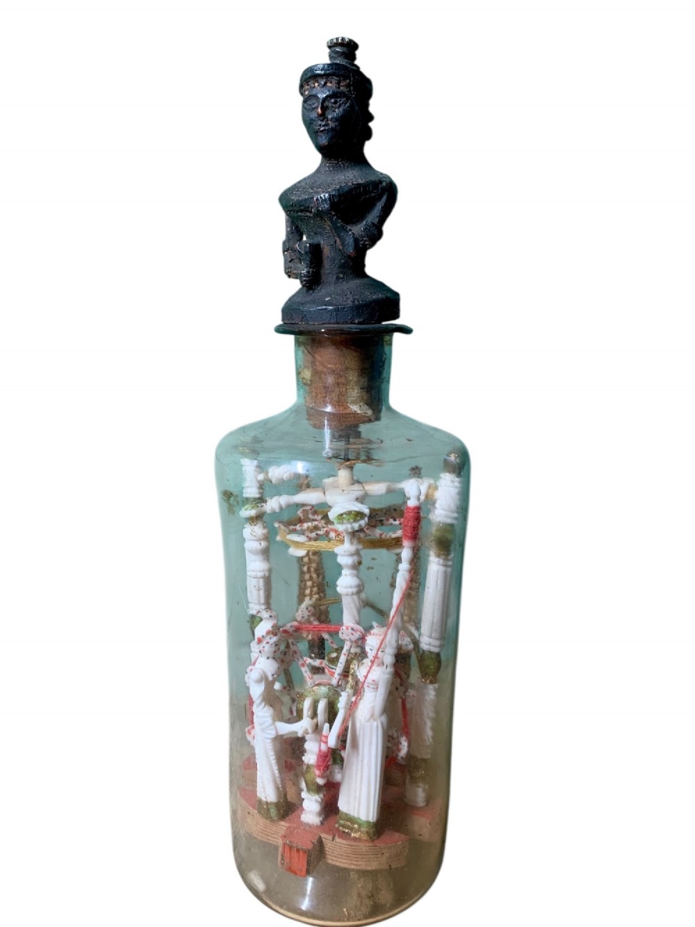 A clear glass bottle housing a detailed hand-carved bone miniature structure inside, topped with a cork and a dark carved bust finial stopper.