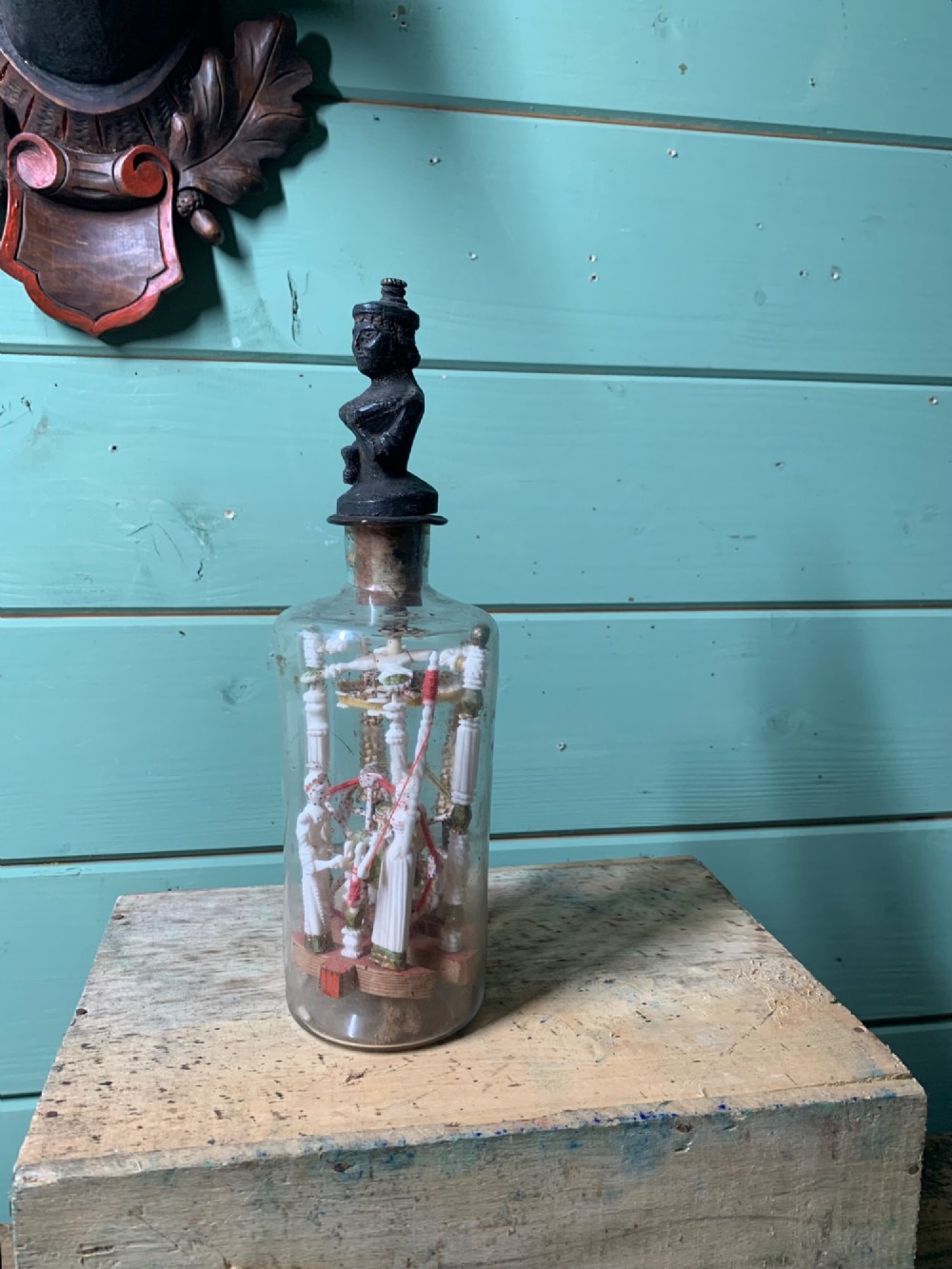 A clear glass bottle housing a detailed hand-carved bone miniature structure inside, topped with a cork and a dark carved bust finial stopper. View II