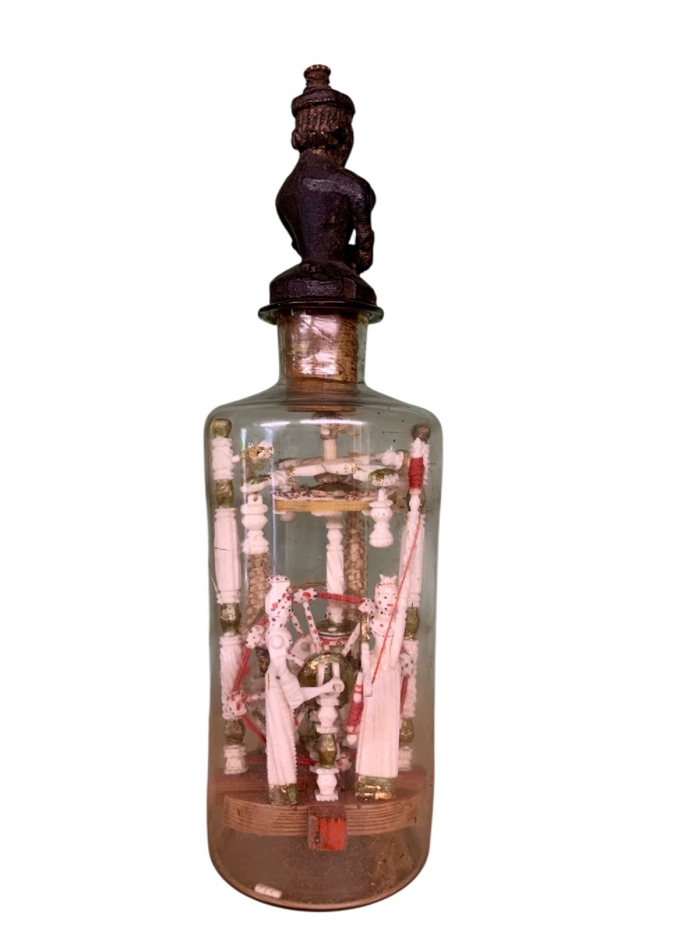A clear glass bottle housing a detailed hand-carved bone miniature structure inside, topped with a cork and a dark carved bust finial stopper. View III