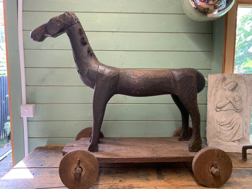 A naive hand-carved dark wood figure of a horse mounted on a flat wooden base with four simple circular wheels. View XV