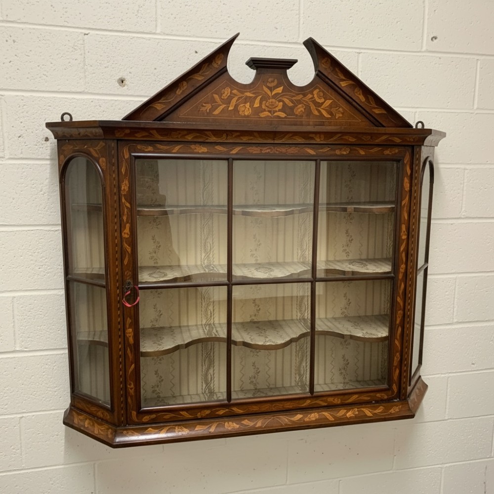 An 18th-century Dutch wall cabinet made of warm brown wood with botanical marquetry inlays, featuring a central glazed door, canted side panels, and a shaped pediment.