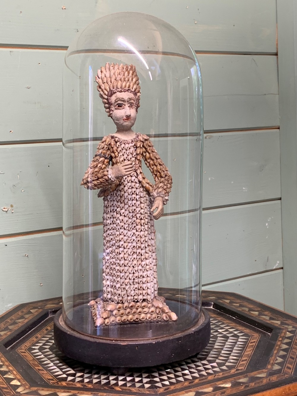 A standing figure of a woman sculpted from tiny arranged seashells on a circular dark wood base.