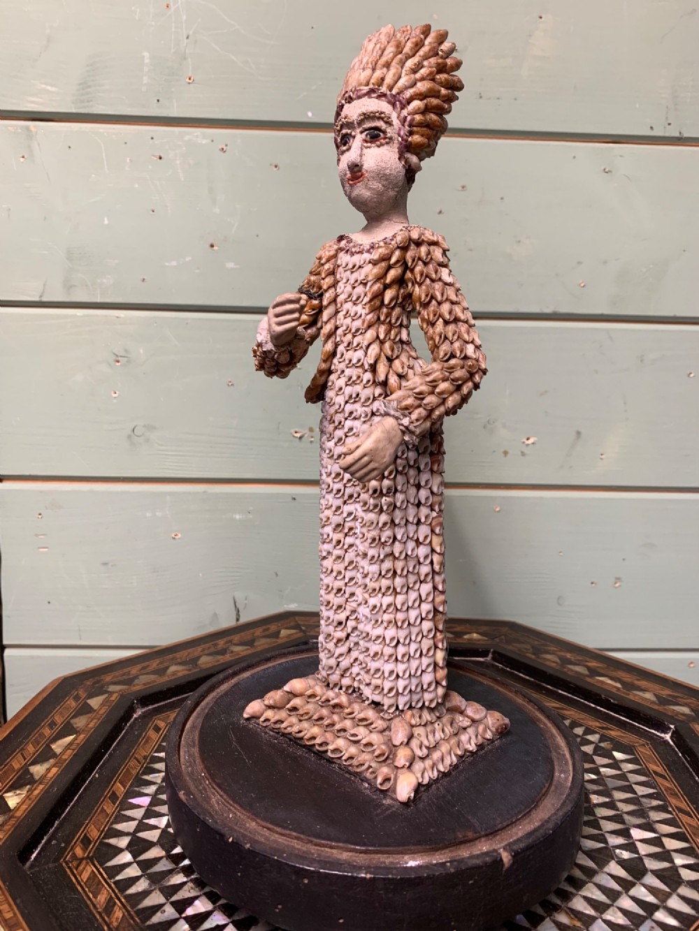A standing figure of a woman sculpted from tiny arranged seashells on a circular dark wood base. View II