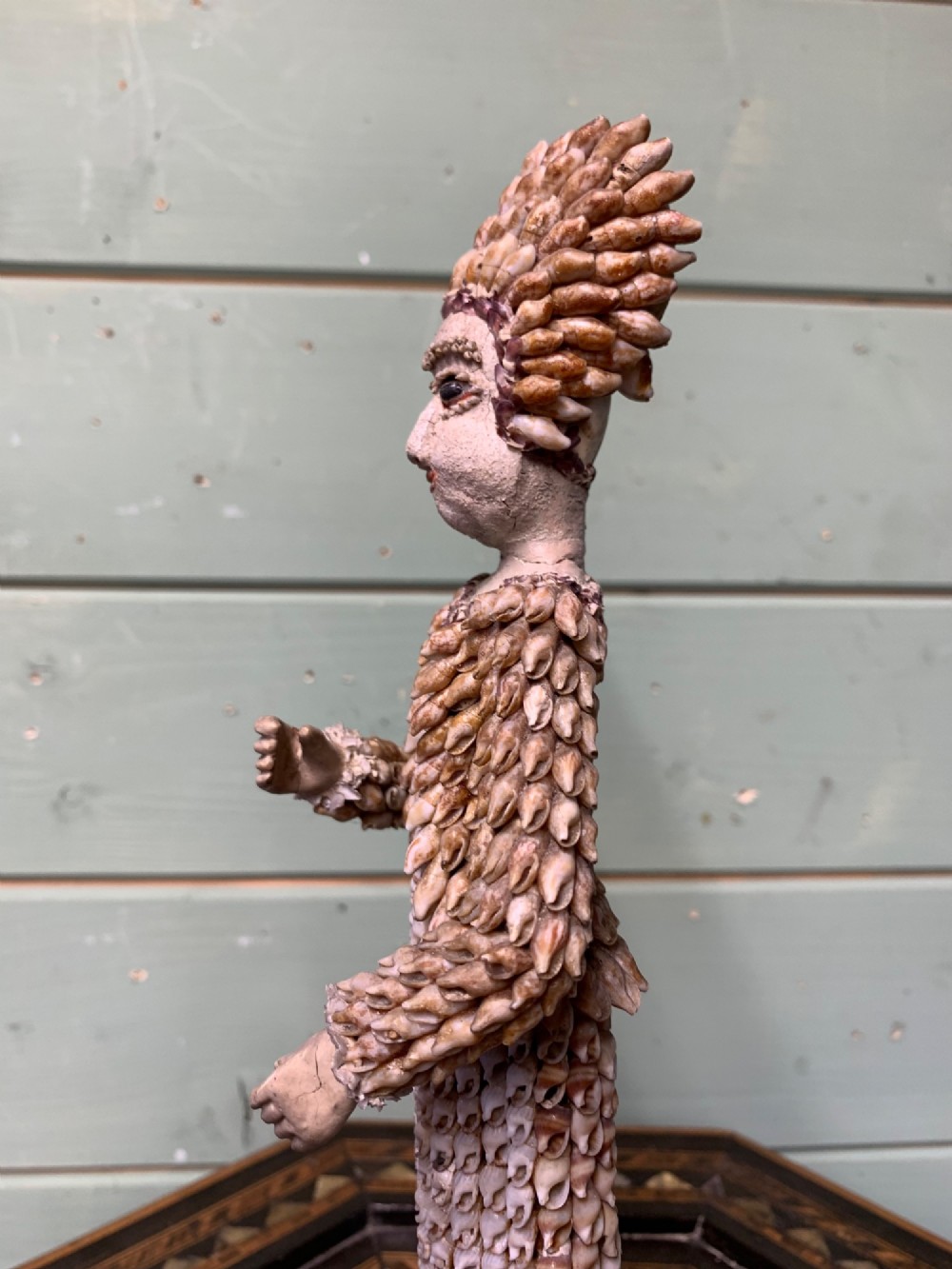 A standing figure of a woman sculpted from tiny arranged seashells on a circular dark wood base. View V