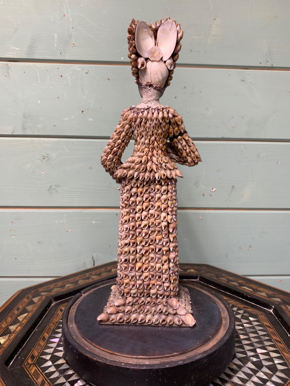 A standing figure of a woman sculpted from tiny arranged seashells on a circular dark wood base. View VIII