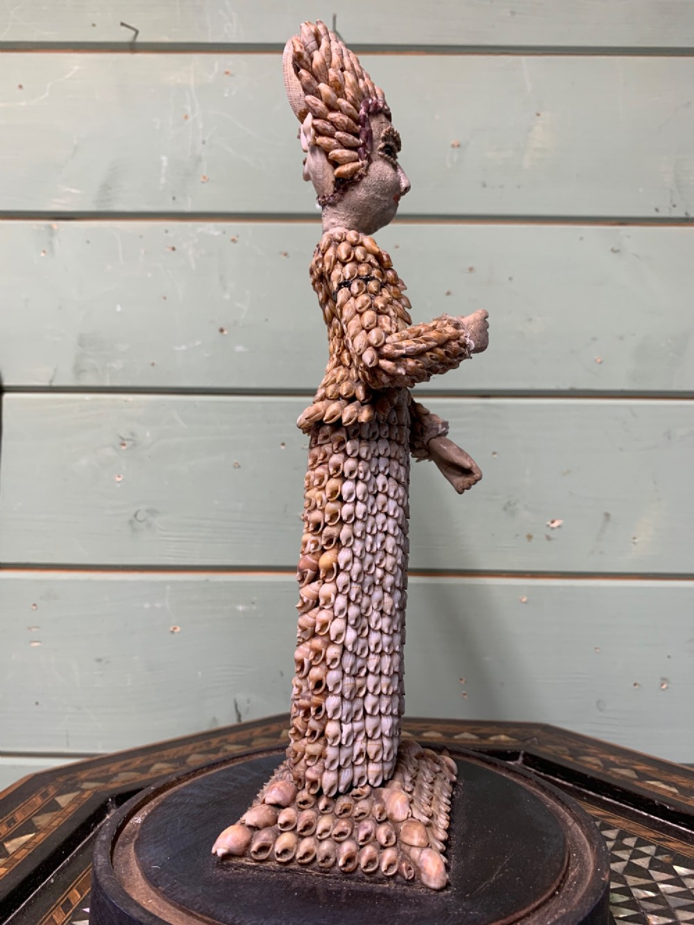 A standing figure of a woman sculpted from tiny arranged seashells on a circular dark wood base. View IX