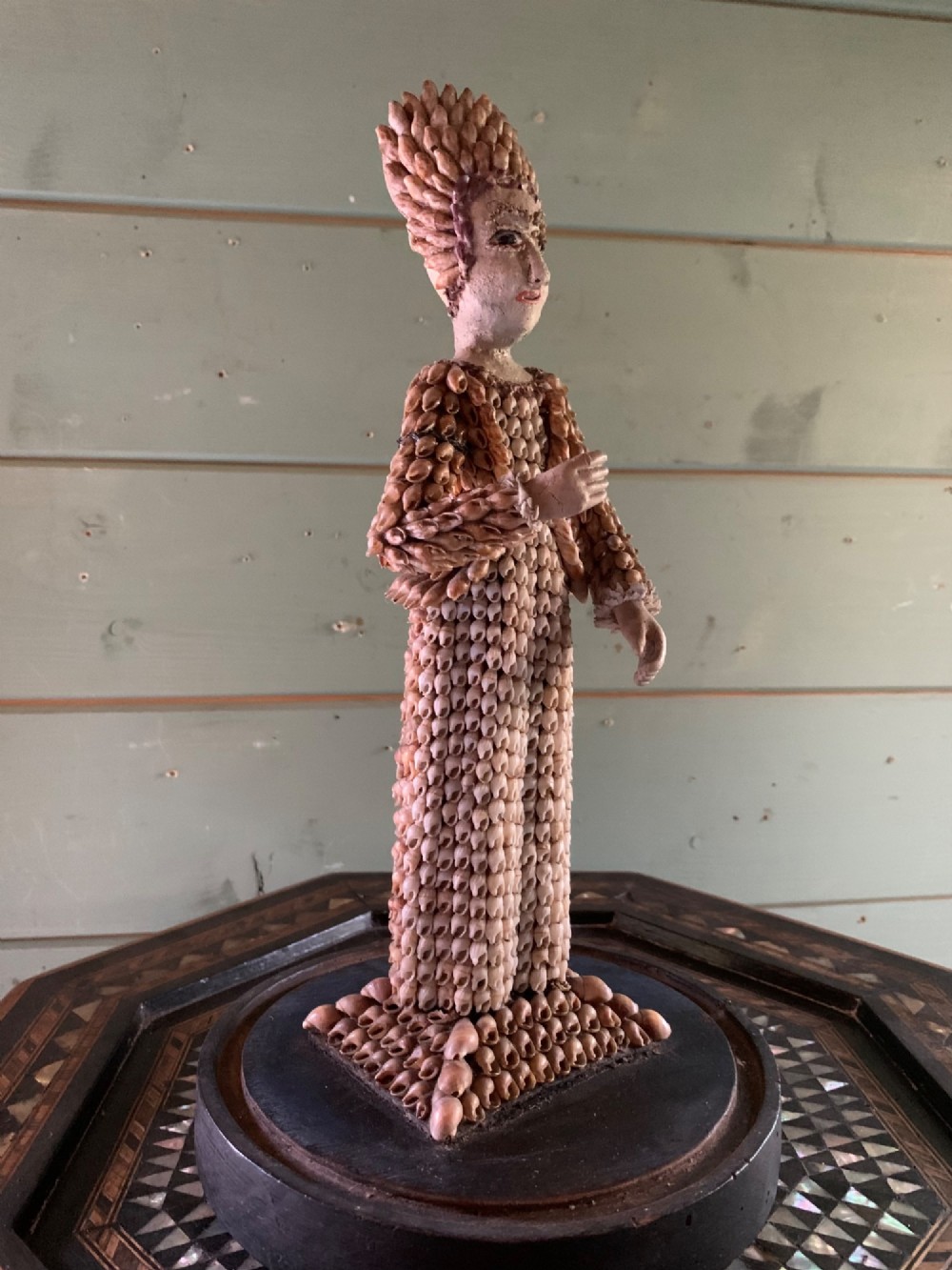 A standing figure of a woman sculpted from tiny arranged seashells on a circular dark wood base. View X